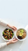 Hands holding two healthy bowls with fresh salad, quinoa, avocado, grilled chicken and black beans on marble surface for nutritious meal planning.