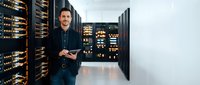 Young Caucasian male IT technician working with tablet in modern server room data center with glowing equipment racks for network maintenance.