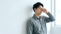 Young Asian man in casual gray shirt leaning against white wall, hand to face in thoughtful pose, suitable for lifestyle, wellness, and mental health content.