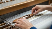 Skilled artisan hands weaving white threads on traditional wooden loom, showcasing ancient textile craftsmanship and handmade fabric production techniques.