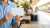 Man in light blue shirt holding blank white business card at outdoor cafe or restaurant with blurred background, suitable for corporate branding and marketing materials.