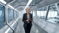 Mature woman with blonde hair, Caucasian ethnicity, wearing business attire and holding a tablet, walking through a modern futuristic corridor with bright natural light and sleek metallic surfaces.