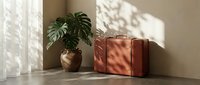 Vintage brown leather suitcase beside a monstera plant in a sunlit minimalist room with warm shadows on beige walls, evoking travel and style.