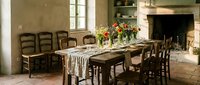 Rustic farmhouse dining room with wooden table, ladder-back chairs, stone fireplace and natural lighting creating warm country home atmosphere.