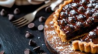 Decadent chocolate ganache tart with sea salt garnish on rustic wooden board, topped with chocolate drizzle and powdered sugar, slice cut to show rich texture.