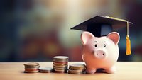Pink ceramic piggy bank wearing graduation cap with gold tassel next to ascending stacks of coins on wooden surface against blurred background, representing education savings.