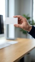 Mature male professional displaying blank white business card in modern office environment with wooden desk and blurred background for branding and corporate identity purposes.