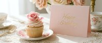 Elegant birthday cupcake with pink rose on floral plate beside a pastel greeting card with gold lettering, soft romantic table setting.
