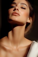 Close-up portrait of young woman with glowing skin, glossy lips and dramatic lighting against dark background. Natural makeup and white clothing create elegant mood.