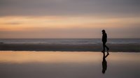 Silhouetted figure jogging along wet beach at sunset, reflected in shallow water. Suitable for fitness, wellness, and lifestyle marketing materials.