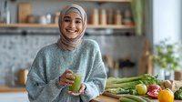 Smiling woman in hijab enjoying healthy lifestyle with green smoothie in modern kitchen surrounded by fresh vegetables and fruits.