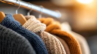 Cozy knitted sweaters hanging on wooden hangers in closet with warm lighting, showcasing different textures and autumn colors in soft focus background.