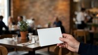 Caucasian male hand holding blank white business card in modern restaurant or cafe setting with blurred background, suitable for corporate branding and networking concepts.
