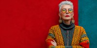 Stylish senior woman with gray hair and yellow glasses wearing colorful knitted sweater against vibrant red and teal wall background.