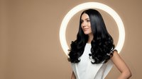 Young Asian woman with long wavy black hair smiling confidently against beige background with circular light frame for beauty and haircare concepts.