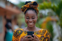 Young African woman in traditional yellow ankara print dress and matching headwrap smiling while using smartphone, perfect for lifestyle, fashion, and mobile technology marketing.