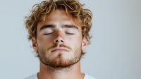 Young Caucasian man with curly blonde hair and beard sleeping peacefully against light background, eyes closed with serene expression showing mindfulness and relaxation.