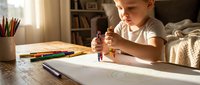 Young blonde child concentrating while drawing with colorful crayons at wooden desk in cozy bedroom with warm natural lighting and art supplies.