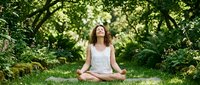 Young woman practicing yoga meditation in peaceful green forest setting with lush foliage and natural sunlight filtering through trees.