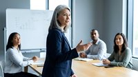 Confident mature Asian businesswoman presenting to diverse team in modern conference room during corporate meeting.