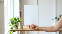 Caucasian hand holding blank white paper or card against wooden shelf with potted green plant in bright modern interior space.