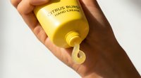 Citrus butter hand cream pouring from yellow bottle held by person with dark skin tone, showcasing product texture and consistency for skincare and cosmetic marketing purposes.
