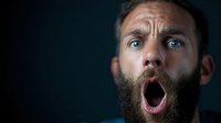 Dramatic portrait of bearded Caucasian man with wide open mouth expressing shock and surprise against dark background, side lighting creates moody atmosphere.