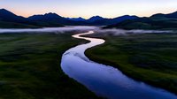 Meandering river through misty valley at dawn, with mountain silhouettes and low-lying fog creating dramatic atmosphere in wilderness landscape. For travel and nature designs.