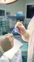 Medical professional in white coat and latex gloves holding laboratory test tube with purple liquid in sterile operating room environment with medical equipment and monitors.