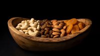 Wooden bowl filled with mixed nuts including cashews, walnuts, almonds and dried apricots on black background for healthy snacking and cooking ingredients.