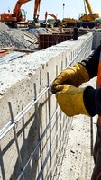 Construction worker in yellow safety gloves securing metal fencing on concrete foundation at active building site with heavy machinery and equipment in background.