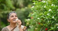 Mature woman with gray hair and South Asian ethnicity, wearing traditional clothing, smiling joyfully while touching white flowers in a lush garden setting. Suitable for wellness, gardening, nature, lifestyle and senior living concepts.