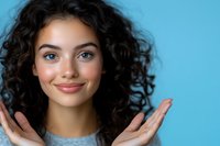 Young woman with curly dark hair and bright smile expressing joy and positivity against blue background. Natural makeup and casual style for authentic lifestyle content.