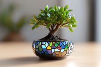 Miniature bonsai tree in decorative mosaic pot with stained glass pattern featuring vibrant blues, greens and yellow colors on wooden surface with soft blur background.