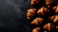 Fresh baked golden croissants cooling on dark wire rack against black background, top view with moody lighting and copy space for menu design.
