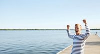 Young child in striped shirt celebrating with arms raised on wooden dock overlooking calm lake water on sunny day, expressing joy and freedom outdoors.