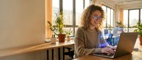 Young mixed-race businesswoman with curly hair and glasses working on laptop in modern bright office space with plants and natural lighting.