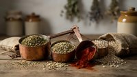 Copper measuring cups filled with green lentils and red paprika powder on rustic wooden table with burlap sack and dried herbs in background.