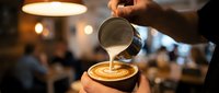 Professional barista creating latte art by pouring steamed milk into coffee cup in modern cafe with warm ambient lighting and blurred background.