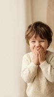Adorable young boy with brown hair covering mouth with hands while laughing in cozy beige sweater against neutral background for family content.