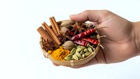 Hand holding wooden bowl with aromatic spices including cinnamon sticks, star anise, cardamom pods, turmeric and chili peppers on white background.