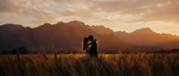 Romantic couple silhouette embracing in golden wheat field at sunset with dramatic mountain landscape backdrop creating intimate wedding engagement scene.