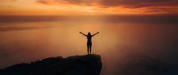 Silhouetted figure with arms raised standing on rocky cliff edge against dramatic golden and orange sunset sky, expressing freedom and achievement.