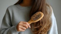 Young woman with long brown hair brushing with wooden hairbrush while wearing cozy gray sweater, self-care routine for healthy hair.