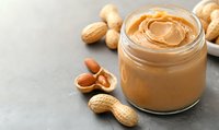 Creamy peanut butter in glass jar with whole and shelled peanuts scattered on gray concrete background, selective focus with copy space for food design.