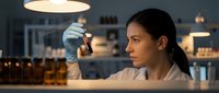 Young woman with dark hair and South Asian ethnicity examining bottle of alcohol at bar counter during evening shift, professional bartender or sommelier inspecting quality of spirits for service.