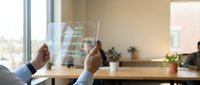 Hands holding transparent digital interface with green data visualization and graphs in modern office workspace with window view and plants.