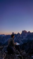 Male hiker reading map on mountain peak during sunset with dramatic alpine landscape and purple twilight sky for adventure travel concepts.