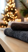 Soft gray towels stacked on wooden surface with blurred Christmas tree lights and candle in background creating cozy holiday atmosphere for spa and wellness concept.