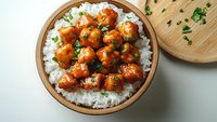 Sweet and sour chicken pieces glazed with Asian sauce served on steamed white rice in wooden bowl, garnished with fresh chopped herbs. Popular takeout dish.